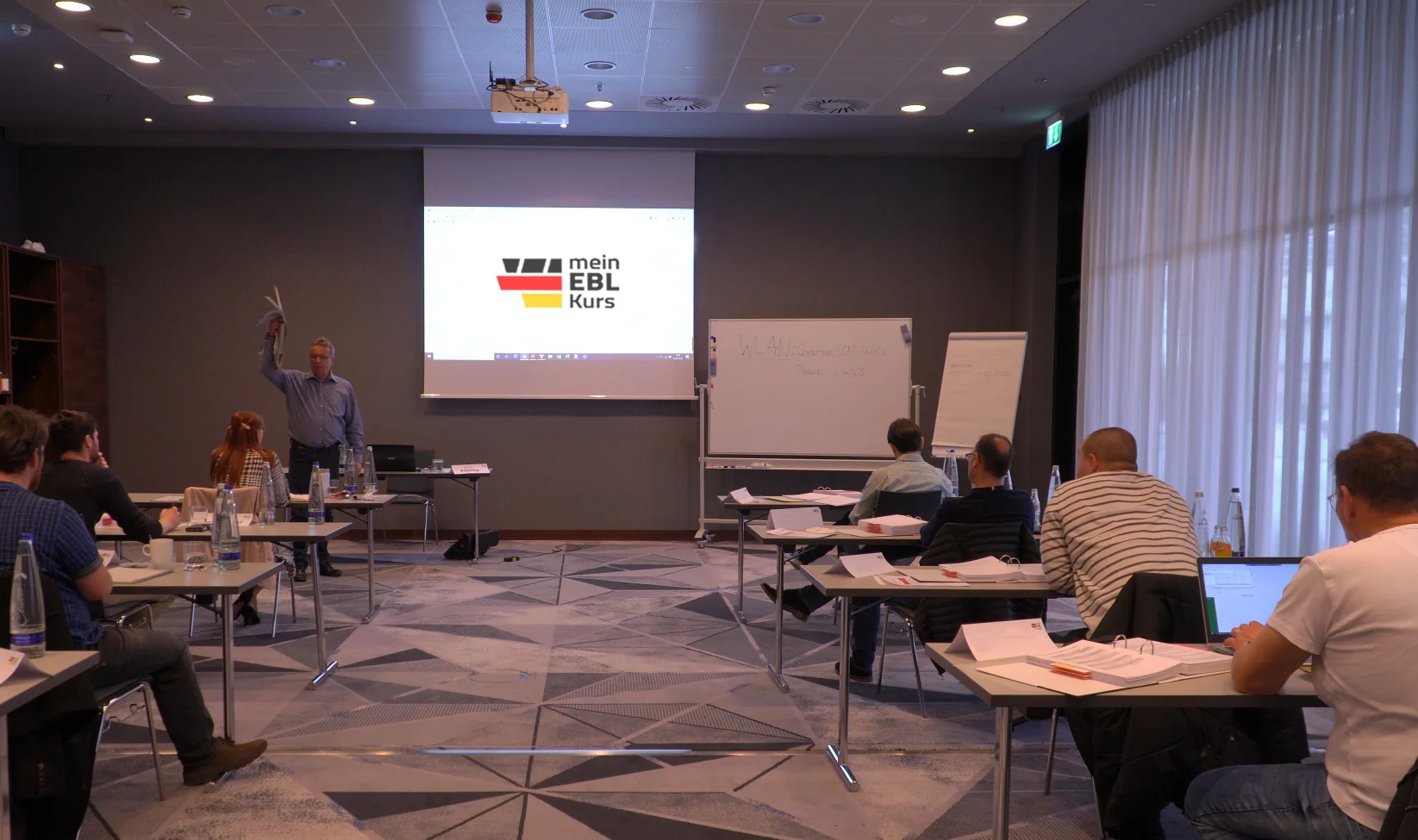 Dozent beim Vortrag im Kursraum mit Logo auf der Leinwand